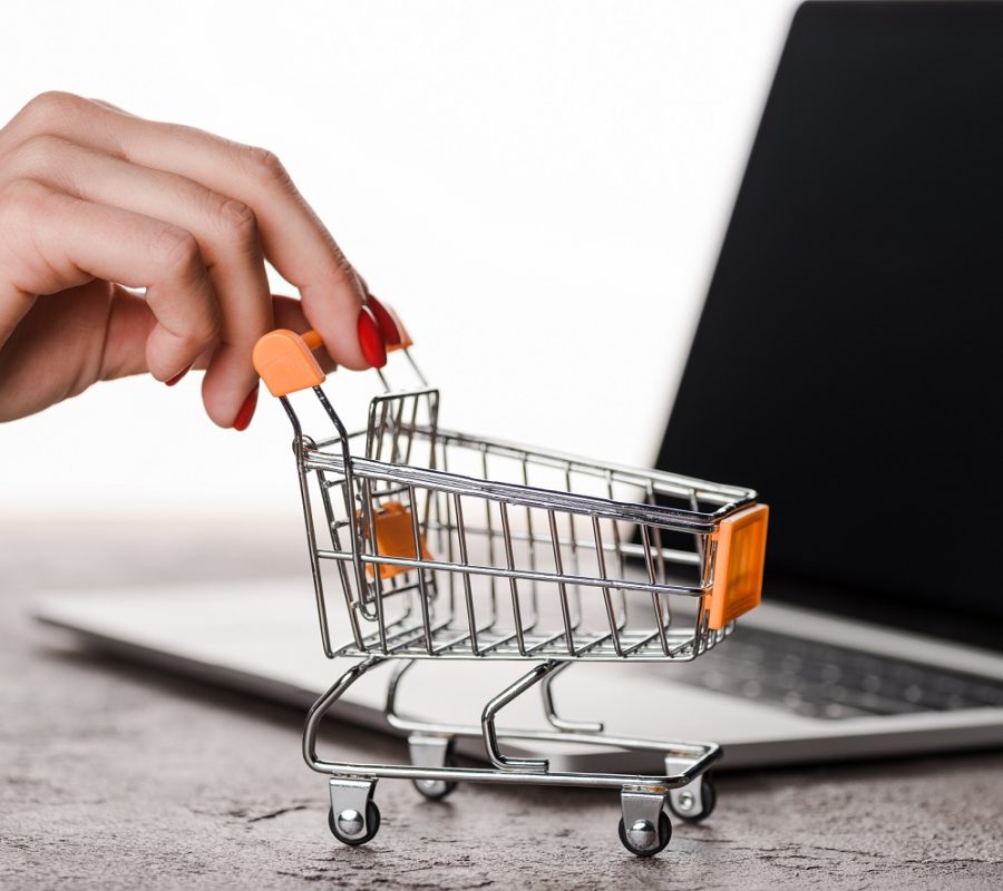 cropped view of woman holding toy shopping cart near laptop isolated on white, e-commerce concept